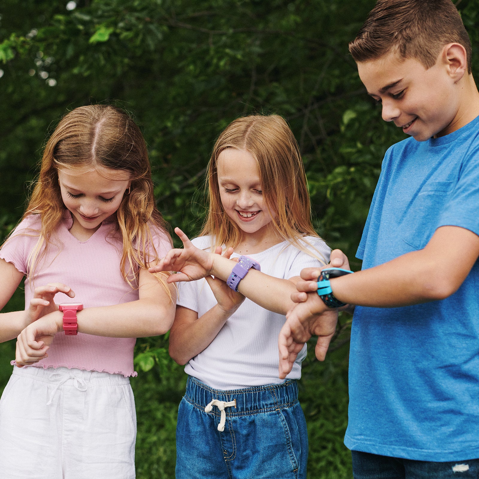 Three children outdoors, looking at smartwatches on their wrists.